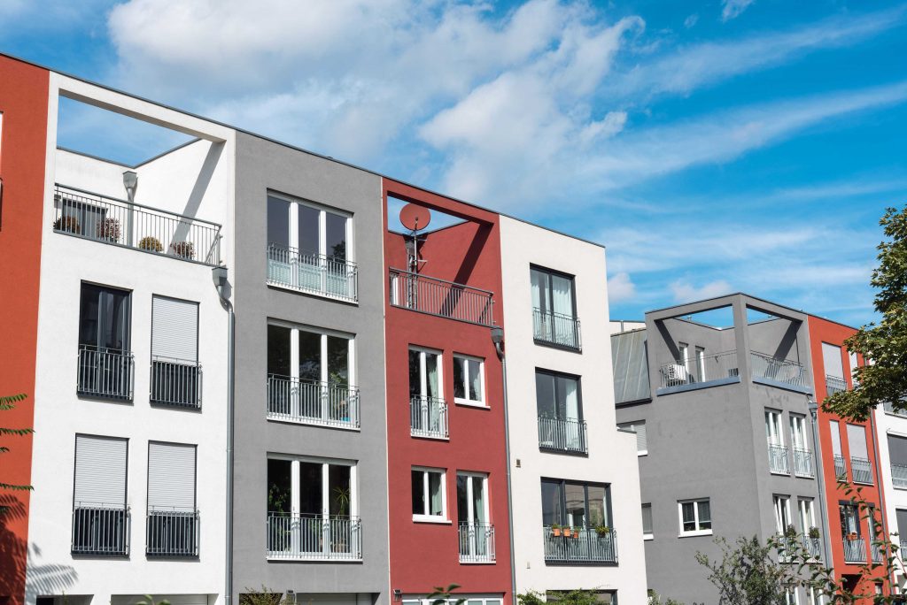 Modern multi-story buildings with red, gray, and white facades, balconies, and a clear blue sky.