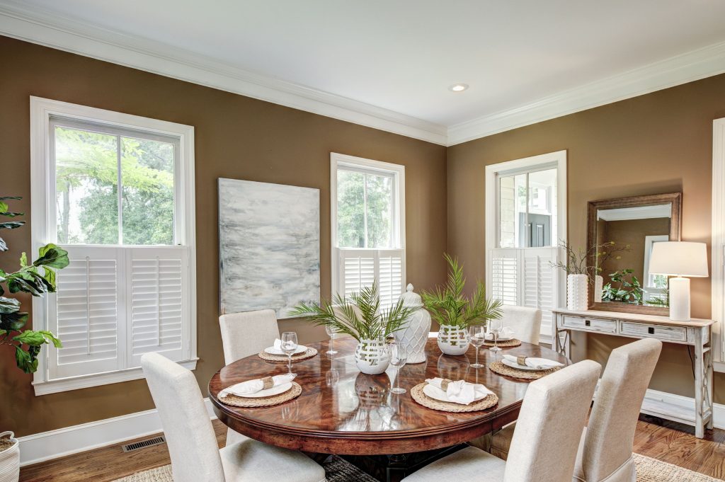 Elegant dining room with a round wooden table, white chairs, plants, artwork, and large windows with shutters.