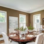 Elegant dining room with a round wooden table, white chairs, plants, artwork, and large windows with shutters.